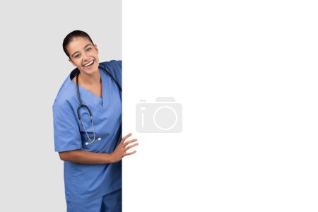 Photo for Friendly nurse in blue scrubs with blank white banner with welcoming smile, perfect for messages, with stethoscope on light background. Sale, medical care, professional advice - Royalty Free Image