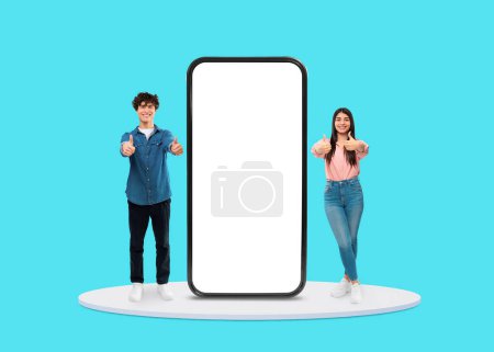 Téléchargez les photos : Jeune homme et femme donnant pouces enthousiastes à côté de l'écran blanc du smartphone, parfait pour la promotion de l'application sur fond bleu, maquette - en image libre de droit
