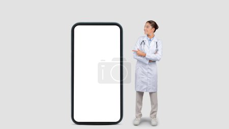 Photo for Happy caucasian millennial doctor in white coat point finger at big phone with empty screen, isolated on gray studio background, collage, panorama. Medical health care app, website recommendation - Royalty Free Image