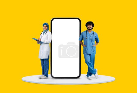 Foto de Doctora con portapapeles y enfermera masculina apoyada en una pantalla masiva de teléfonos inteligentes, que representa la atención médica moderna, sobre un fondo amarillo - Imagen libre de derechos