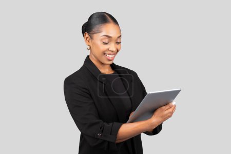 Foto de Una joven empresaria afroamericana enfocada en un traje negro utiliza una tableta, posiblemente revisando documentos importantes o realizando negocios en línea, aislada en un fondo de estudio gris - Imagen libre de derechos
