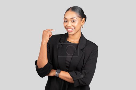 Foto de Joven mujer afroamericana alegre en una chaqueta negra, reloj inteligente en la muñeca, haciendo un gesto fuerte con una sonrisa, simbolizando la confianza y el éxito, aislado en el fondo gris del estudio - Imagen libre de derechos