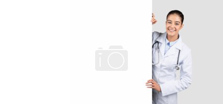 Téléchargez les photos : Heureuse femme médecin caucasienne millénaire en manteau blanc avec une énorme bannière avec espace libre, isolée sur fond de studio gris, panorama. Présentation médicale, soins de santé, vente, publicité d'attention et offre - en image libre de droit
