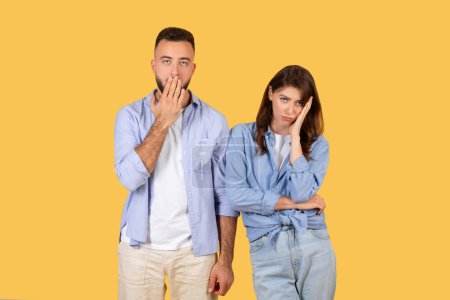 Photo for Remorseful man places his hand over his mouth while woman rests her cheek on her hand, both showing signs of regret or worry on yellow background - Royalty Free Image