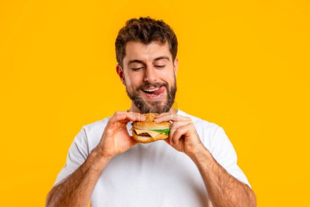 Photo for Cheat Meal. Portrait Of Funny Hungry European Man Eating Tasty Cheeseburger, Standing Against Yellow Background In Studio. Junk Food Nutrition, Concept Of Unhealthy Meals - Royalty Free Image