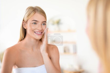 Photo for Happy blonde lady indulges in skincare ritual, softly pressing cotton pad against her skin in contemporary bathroom. Woman applying moisturizing lotion near mirror. Selective focus - Royalty Free Image