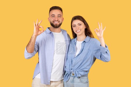 Photo for Happy man and woman in casual blue attire making OK gestures with their hands, signaling approval and satisfaction on cheerful yellow backdrop - Royalty Free Image