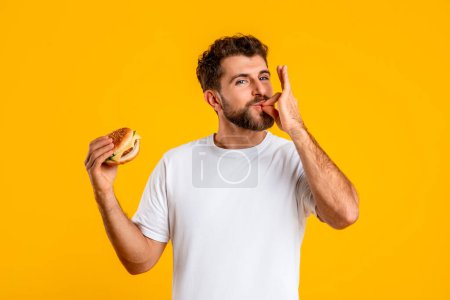 Photo for Delicious Fastfood. Portrait Of Smiling Hungry Man Eating Burger And Licking Fingers, Standing Over Yellow Studio Wall. Happy Casual Guy Man Holding Snack Looking At Camera With Smile - Royalty Free Image