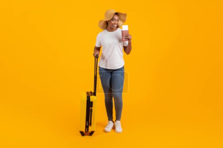 Téléchargez les photos : Heureuse femme afro-américaine millénaire en pose décontractée avec valise et billets de carte d'embarquement, portant un chapeau d'été prêt pour les vacances, fond de studio jaune, pleine longueur, espace de copie - en image libre de droit