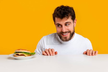 Foto de Divertido tipo hambriento mirando sabrosa hamburguesa en el escritorio posando en el estudio sobre fondo amarillo, teniendo antojos de comida y el hábito de comer en exceso. Concepto de comida engañosa, dieta y nutrición - Imagen libre de derechos
