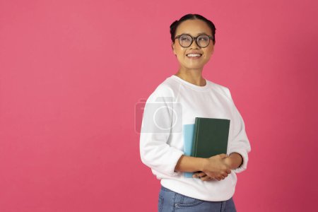 Téléchargez les photos : Éducation académique. Souriante jeune femme asiatique portant des lunettes de vue tenant des livres et regardant l'espace de copie, heureuse étudiante coréenne debout isolé sur fond de studio rose, profiter de l'apprentissage - en image libre de droit