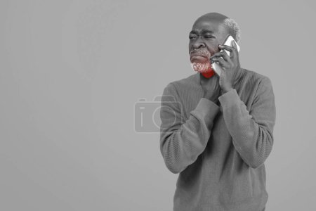 Photo for Distressed senior man in a grey sweater grimacing in pain while talking on a white phone against a monochromatic background, depicting a problematic conversation, flu and cold - Royalty Free Image