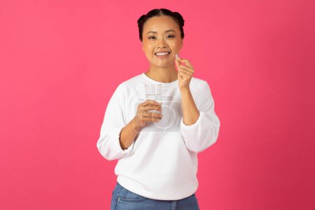 Foto de Feliz joven asiática dama sosteniendo vaso de agua y belleza suplemento píldora, milenaria sonriente mujer tomando cápsula de vitamina para una inmunidad saludable o piel hermosa, de pie aislado sobre fondo rosa - Imagen libre de derechos