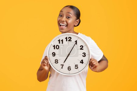 Foto de Es la hora. Joven mujer afroamericana alegre mostrando reloj redondo sonriendo a la cámara, mostrando el concepto de plazo y cosquillas segundos sobre el fondo del estudio amarillo. Gestión del tiempo - Imagen libre de derechos