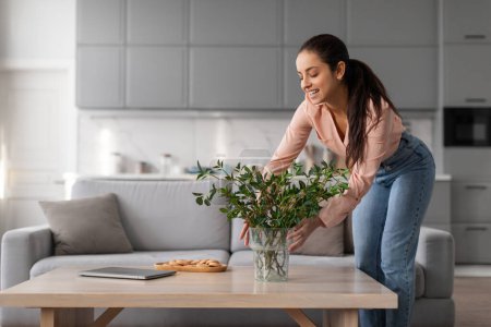 Photo for Smiling woman places plant in vase on the center of coffee table, enhancing the rooms natural aesthetics in her bright living space, free space - Royalty Free Image