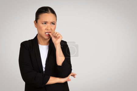 Photo for Worried Asian businesswoman biting her fingernail with a nervous expression, showing anxiety or concern, dressed in a professional black blazer over a white shirt on a light grey background - Royalty Free Image