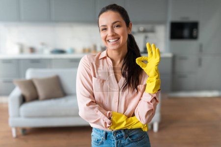 Téléchargez les photos : Femme rayonnante en chemise pastel et en denim, portant des gants de nettoyage jaunes, des poutres à la caméra, prête à commencer sa routine de nettoyage de printemps dans une maison rangée - en image libre de droit