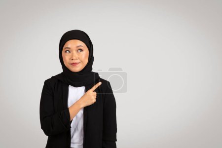 Photo for A contemplative young Asian businesswoman in a hijab points to the side with an expressive look, suggesting an idea or direction in a corporate setting, isolated on gray background, studio - Royalty Free Image