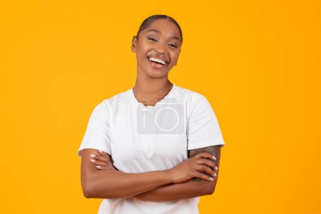Téléchargez les photos : Portrait de femme noire heureuse en t-shirt blanc croisant les mains et posant avec confiance, offrant un sourire joyeux sur fond de studio jaune, incarnant le style féminin confiant et la beauté - en image libre de droit