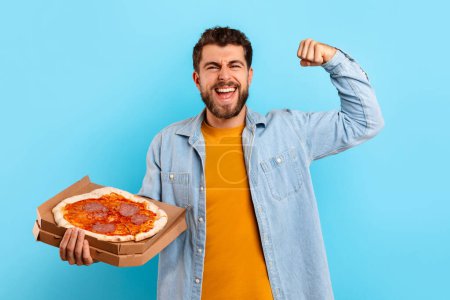 Téléchargez les photos : Amoureux de la malbouffe. Joyeux gars tenant une boîte à pizza et montrant ses biceps muscles levant le bras, célébrant le jour du repas Cheat, souriant à la caméra sur fond de studio bleu. Dépendance nutritionnelle malsaine - en image libre de droit