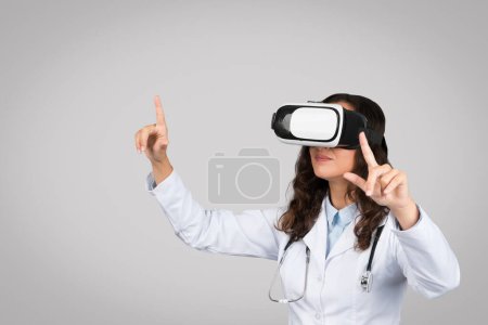 Téléchargez les photos : Dispositif d'éducation médicale. Médecine en manteau avec stéthoscope expérimentant la réalité virtuelle en lunettes vr, portant casque et geste, isolée sur fond de studio gris - en image libre de droit