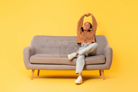 Foto de Relajada adolescente afroamericana estirando los brazos mientras está sentada en el sofá, mostrando comodidad y relajación en traje casual contra el fondo del estudio amarillo. Longitud completa, espacio de copia - Imagen libre de derechos