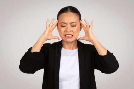 Photo for Stressed Asian businesswoman with a pained expression, holding her head in frustration or headache, dressed in a professional black blazer and white shirt, against a grey background - Royalty Free Image