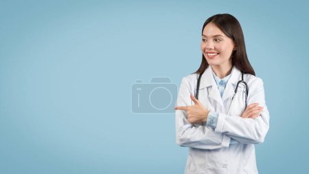 Téléchargez les photos : Jeune femme médecin souriante avec stéthoscope autour du cou, pointant avec confiance vers son côté à l'espace libre sur fond bleu apaisant, panorama - en image libre de droit