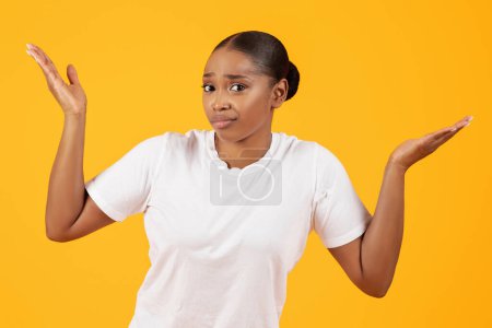 Foto de No lo sé. Unsure Puzzled Black Woman Shrugging Shoulders Standing Posing Against Yellow Studio Backdrop, Looking At Camera With Doubtful Expression (en inglés). Concepto de gesto de encogimiento de hombros. - Imagen libre de derechos