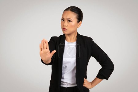 Foto de Empresaria asiática asertiva en una chaqueta negra con una expresión seria haciendo un gesto de stop con la mano, señalando límite o rechazo, sobre un fondo gris. Negocios y trabajo - Imagen libre de derechos