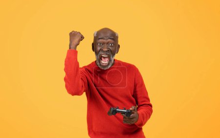 Photo for Excited elderly African American man with a beard wearing a red sweater triumphantly raises his fist while holding a video game controller, with a victorious yell on a yellow background - Royalty Free Image