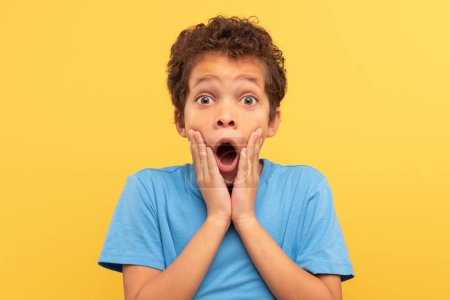 Photo for Shocked boy with curly hair, hands on cheeks and mouth wide open in astonishment, wearing blue shirt against yellow background, perfectly capturing a moment of surprise - Royalty Free Image