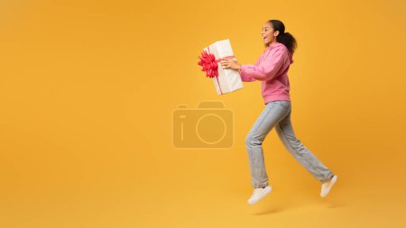 Téléchargez les photos : Livraison de cadeau d'anniversaire. Joyeux jeune femme afro-américaine offrant boîte cadeau enveloppé, étirant les bras à l'espace libre, posant en plein air sur fond de studio jaune, panorama. Offre vacances - en image libre de droit