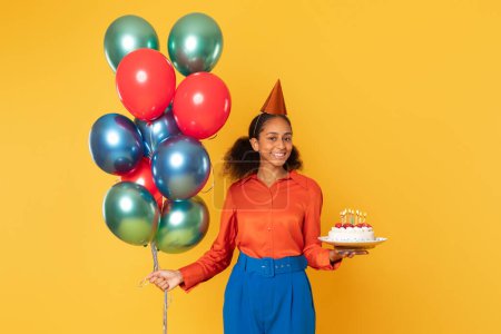 Foto de Chica alegre adolescente negro posando con un montón de globos festivos y pastel de cumpleaños, encarnando la celebración contra el fondo del estudio amarillo, sonriendo a la cámara. Bday vacaciones y diversión de fiesta - Imagen libre de derechos