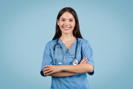Photo for Young caucasian woman nurse in blue coat stands with folded arms, smiling warmly against blue background, exuding professionalism and care - Royalty Free Image