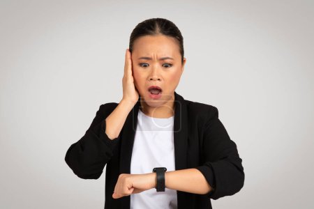 Foto de Una mujer de negocios asiática sorprendida comprueba el tiempo en su reloj inteligente con una mano en la mejilla, retratando un sentido de urgencia, gestión del tiempo, o estrés plazo, aislado en el fondo gris, estudio - Imagen libre de derechos