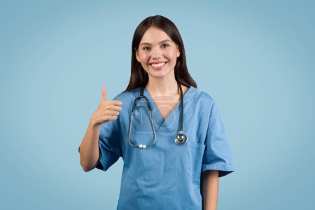 Téléchargez les photos : Infirmière joyeuse, vêtue d'un manteau bleu et portant un stéthoscope, souriante et montrant les pouces vers le haut geste, sur fond bleu - en image libre de droit