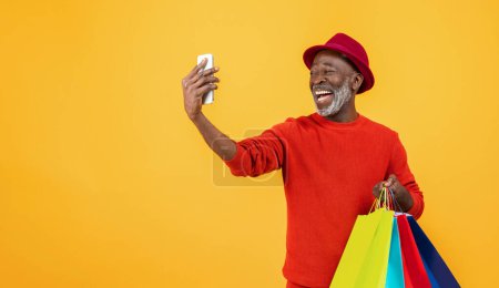 Photo for Smiling senior Black man in a red sweater and hat taking a selfie with a phone, while happily holding a bunch of bright shopping bags on a sunny yellow studio background. - Royalty Free Image