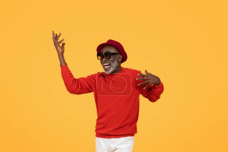 Téléchargez les photos : Homme âgé souriant et exubérant dans un pull rouge et un chapeau, dansant les bras levés, portant des lunettes de soleil, incarnant la joie et des vibrations insouciantes, amusant, sur un fond jaune vif, studio - en image libre de droit
