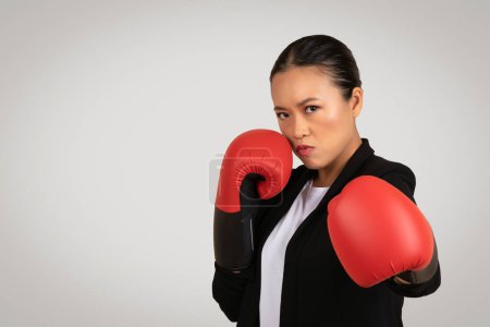 Téléchargez les photos : Femme d'affaires asiatique déterminée en tenue professionnelle avec des gants de boxe est prêt à se battre, symbolisant l'autonomisation, défi, et esprit d'affaires concurrentiel, isolé sur fond gris, studio - en image libre de droit