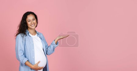 Téléchargez les photos : Mignon attrayant jeune femme attendant étreignant son gros ventre et pointant vers l'espace de copie pour la publicité, isolé sur fond de studio rose, panorama. Grossesse, accouchement, maternité - en image libre de droit