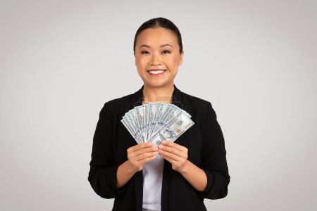 Photo for A radiant Asian businesswoman in professional attire beams as she offers a fan of hundred-dollar bills, representing financial success and economic transactions, isolated on gray background, studio - Royalty Free Image