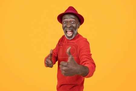 Foto de Hombre afroamericano senior extático en un suéter rojo y sombrero dando un pulgar hacia arriba, su cara radiante de felicidad y entusiasmo contra un estudio de fondo amarillo brillante - Imagen libre de derechos