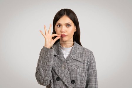 Photo for Professional young woman in grey plaid blazer making silence gesture with her finger on her lips, signaling the need for quiet or secrecy - Royalty Free Image