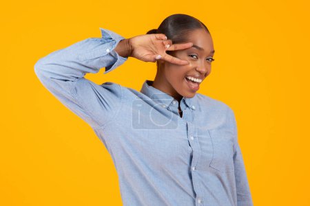 Photo for Joyful Black Young Woman In Casual Denim Shirt Having Fun Gesturing Victory Sign Near Face And Smiling To Camera Over Yellow Studio Backdrop. Good Vibes And Positive Emotions Concept - Royalty Free Image