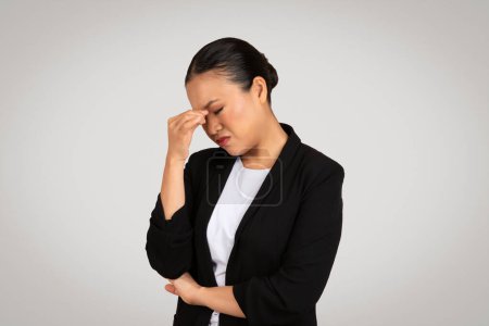 Foto de Mujer de negocios asiática preocupada con chaqueta de traje negro tocando su sien, mostrando un gesto de estrés, dolor de cabeza o pensamiento profundo, con una expresión problemática sobre un fondo gris - Imagen libre de derechos