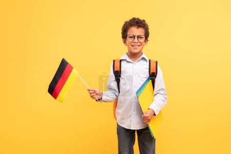 Foto de Feliz colegial con el pelo rizado y gafas, orgullosamente sosteniendo la bandera alemana, con carpetas coloridas de la escuela y la mochila, de pie sobre el fondo amarillo, simbolizando el entusiasmo educativo - Imagen libre de derechos