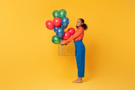 Téléchargez les photos : Adolescente noire positive en tenue lumineuse tenant un tas de ballons festifs colorés dans les mains, célébrant les vacances et présentant des ballons comme cadeau sur fond de studio jaune. Plan complet - en image libre de droit