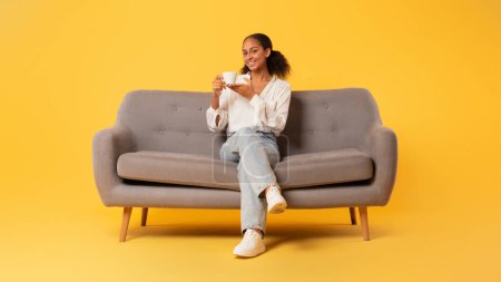 Foto de Chica adolescente negro feliz con taza de café caliente sentado en un cómodo sofá con fondo de estudio amarillo, disfrutando de una bebida sabrosa mientras descansa solo. Largura completa, panorama - Imagen libre de derechos