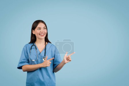 Téléchargez les photos : Jeune infirmière professionnelle en manteau bleu fait des gestes sur le côté à l'espace vide pour la publicité, sur fond bleu vif. Idéal pour les promotions médicales - en image libre de droit